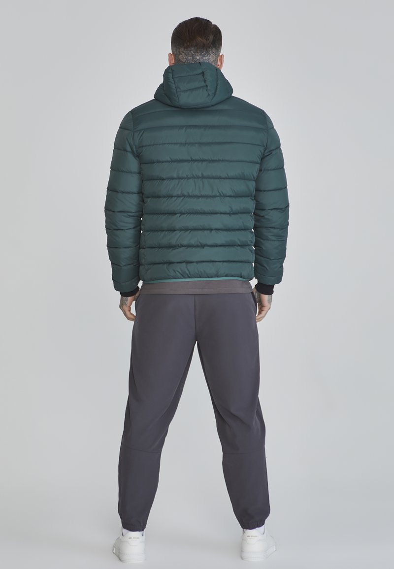 Giacca imbottita verde scuro con cappuccio, caratterizzata da una trama trapuntata e polsini elastici, abbinata a pantaloni della tuta grigi e sneakers bianche.