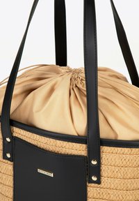 Woven straw tote bag with a beige drawstring fabric top, black leather straps, and a black accent panel featuring a gold logo.