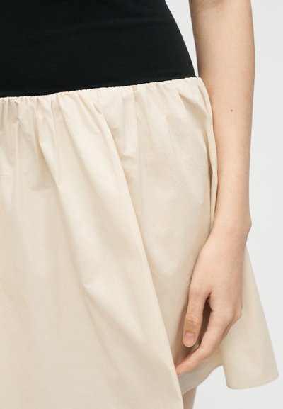 Άτομο που φοράει μαύρο τοπ και μπεζ φούστα, με κοντινή λήψη στη σφικτή μέση της φούστας και το δεξί τους χέρι να ακουμπάει δίπλα τους.