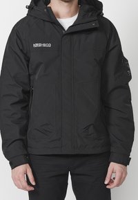 Svart huva jacka med framdragkedja, två stora sidofickor och broderad logotyp. Tillverkad av ett slätt, väderbeständigt material.