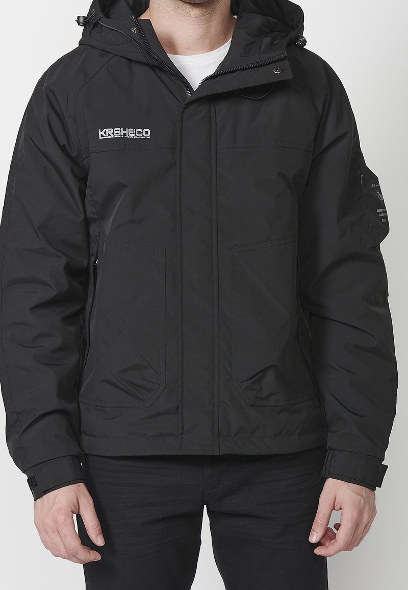 Svart huva jacka med framdragkedja, två stora sidofickor och broderad logotyp. Tillverkad av ett slätt, väderbeständigt material.