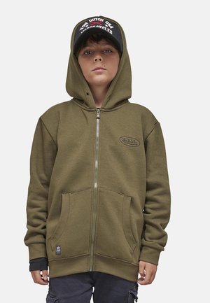 Garçon portant un sweat à capuche zippé vert olive et une casquette noire avec un texte blanc et rouge, debout devant un fond uni clair.