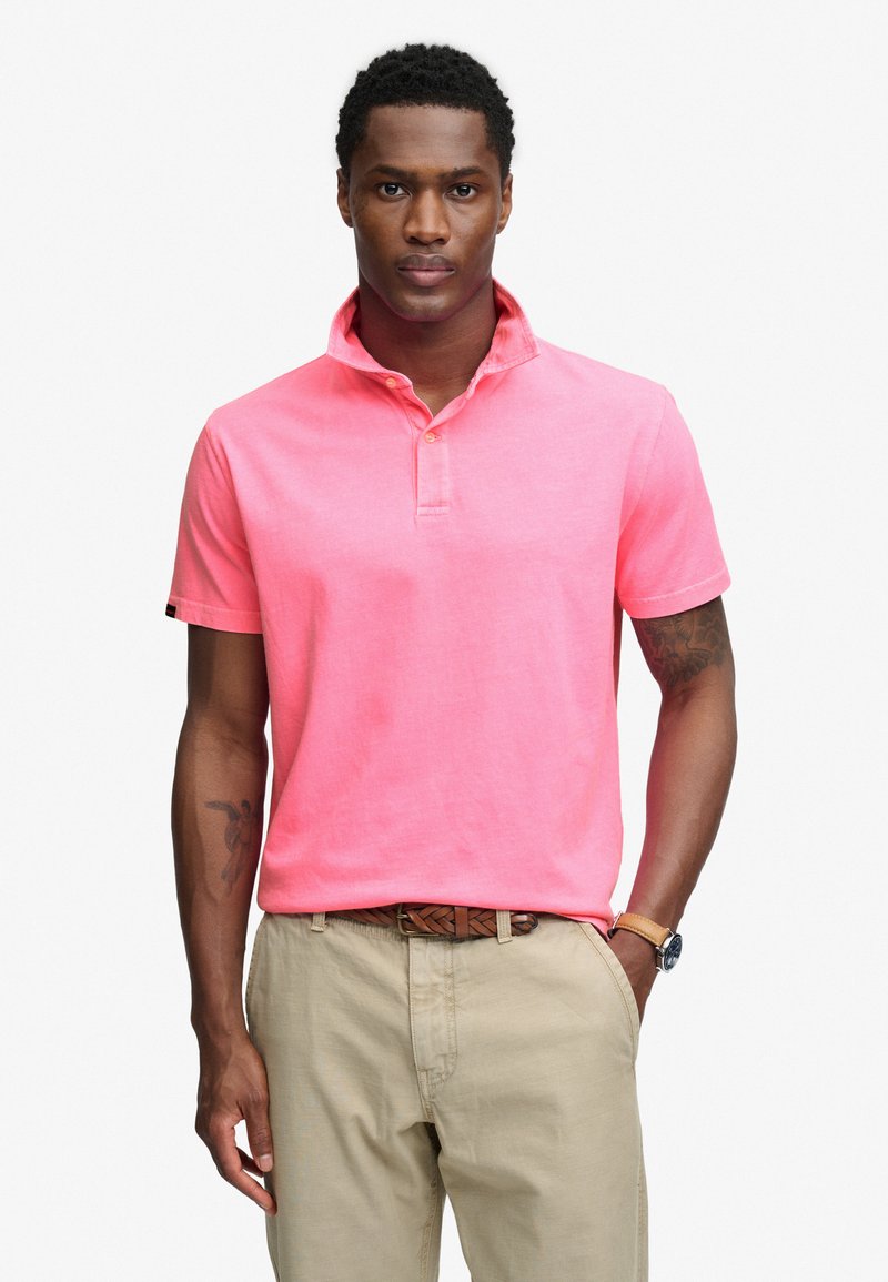 Man in roze poloshirt met korte mouwen en opstaande kraag, beige broek, bruine gevlochten riem, polshorloge, en hand in zak.