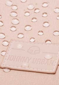 Tessuto rosa resistente all'acqua con gocce d'acqua, caratterizzato da un'etichetta beige testurizzata in rilievo con la scritta "JOHNNY URBAN" e accenti circolari.