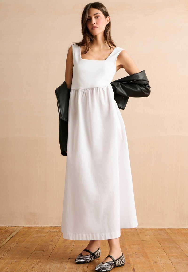 Witte, mouwloze maxi-jurk met een vierkante halslijn en geplooide taille. Gecombineerd met een zwart leren jack en zwart-gepatenteerde ballerina's.