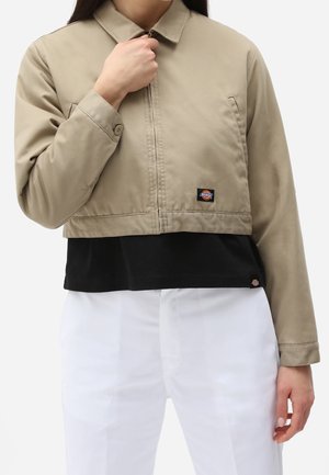Person iført en cropped beige jakke over en sort skjorte og hvide bukser, holder jakke-lynlåsen tæt ved kraven.
