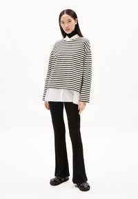 Pullover oversize a righe bianche e nere indossato sopra una camicia bianca, abbinato a pantaloni neri a campana e sandali neri. Tessuto dalla superficie liscia.