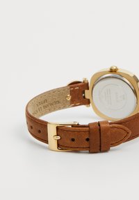 Brauner Lederarmband mit goldener Schnalle, befestigt am goldenen und silbernen Uhrengehäuse auf der Rückseite, das Marken- und Materialdetails zeigt.