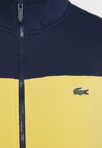 Vêtement zippé bleu marine et jaune avec le logo crocodile Lacoste brodé sur la partie jaune près de la poitrine.