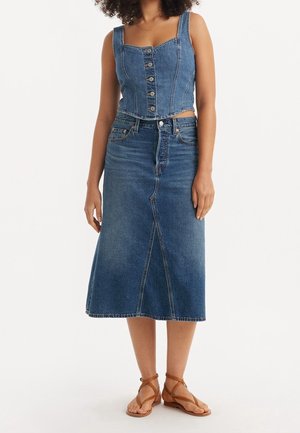 Kobieta ubrana w niebieską, zapinaną na guziki krótka bluzkę z denimu, niebieską spódnicę midi z denimu o wysokim stanę z przednim szwem oraz brązowe, płaskie sandały na paski.