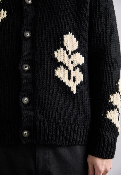 Sort strikket cardigan med store cremede diamantmønstre på ærmerne, med seks sorte knapper samt ribbede ender og manchetter.