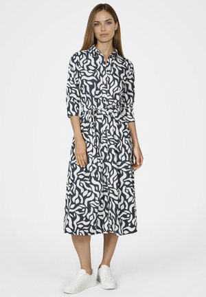 Frau trägt ein midi Hemdblusenkleid mit schwarz-weißem abstraktem Muster, Dreiviertelärmeln und weißen Sneakern vor einem einfarbigen Hintergrund.