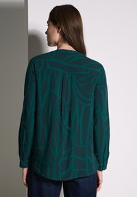 Camicia a maniche lunghe di un verde scuro con un motivo a foglia testurizzato. Il retro presenta una piega per una maggiore libertà di movimento. Vestibilità aderente con polsini abbottonati.