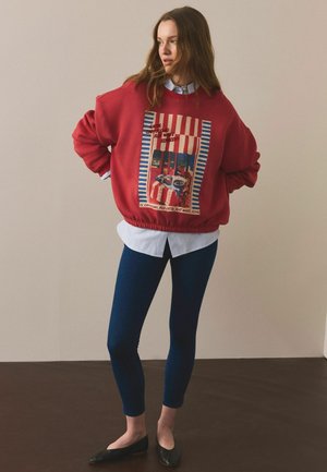 Žena stojaca v interiéri, oblečená v červenej nadrozmernej mikine s farebnou potlačou kaviarne, bielej košeli s golierom, tmavomodrých legínach a čiernych balerínach.