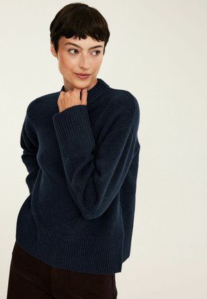 Femme aux cheveux courts et foncés portant un pull en tricot bleu foncé, posant avec la main près du menton et regardant sur le côté devant un fond uni.