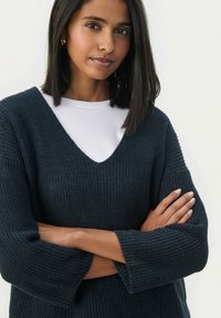 Pull en tricot navy foncé, avec un col en V, des manches larges et un tissu texturé. En dessous, un haut blanc est visible au niveau du col.