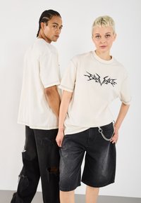 Dva modely stojí zády k sobě a mají na sobě volné béžové trička s černými grafickými potisky, v kombinaci s roztrhanými černými džíny kraťasy.