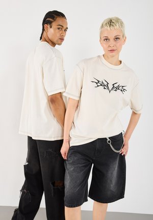 Zwei Models stehen Rücken an Rücken und tragen lässige beige T-Shirts mit schwarzen grafischen Drucken, kombiniert mit verwaschenen schwarzen Jeansshorts.