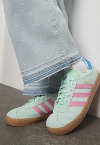 Person med lyseblå jeans og mintgrønne Adidas Gazelle-sneakers med rosa striper og gummisåler, stående på grått gulv.