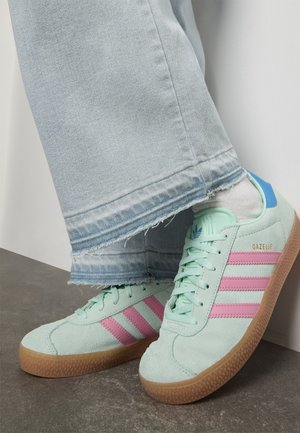 Persoon in lichtblauwe jeans en mintgroene Adidas Gazelle sneakers met roze strepen en rubberen zolen, staande op een grijze vloer.