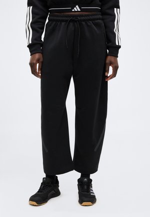 Osoba nosící černé široké tepláky s šňůrkou v pase, černé tenisky Adidas s gumovou podrážkou a černý crop top Adidas s bílými pruhy.