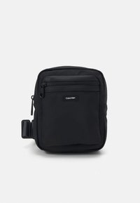 Borsa a tracolla nera in tessuto con una superficie texturizzata, tasca anteriore con zip e un'etichetta con logo. Presenta una clip laterale per un attacco versatile.