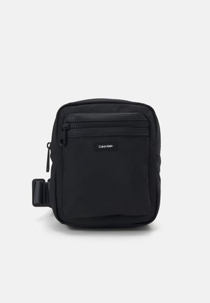 Borsa a tracolla nera in tessuto con una superficie texturizzata, tasca anteriore con zip e un'etichetta con logo. Presenta una clip laterale per un attacco versatile.