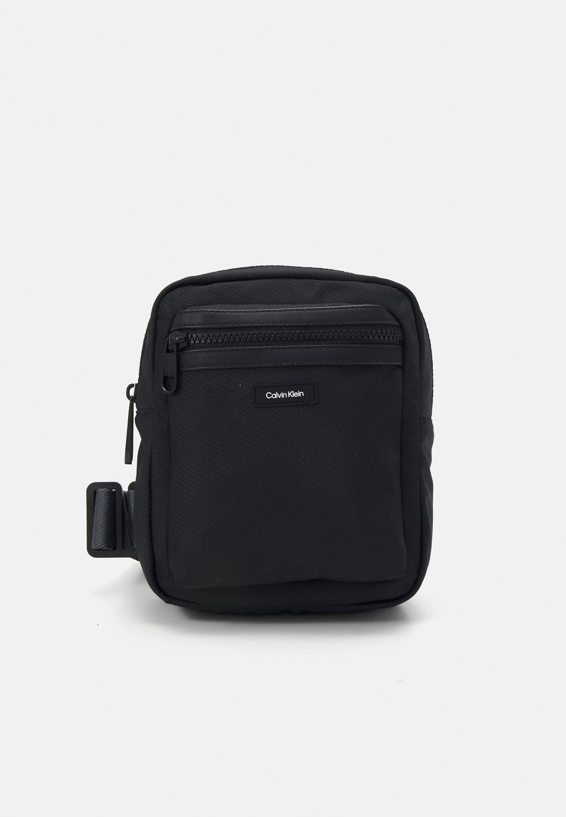 Borsa a tracolla nera in tessuto con una superficie texturizzata, tasca anteriore con zip e un'etichetta con logo. Presenta una clip laterale per un attacco versatile.