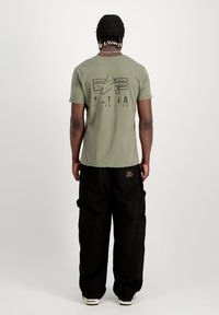 Olivgrön T-shirt med grafisk tryck och logotyp på baksidan, kombinerad med svarta cargobyxor med flera fickor.