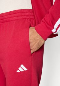 Κόκκινο βαμβακερό παντελόνι jogging με ελαστική μέση, με λευκό λογότυπο Adidas στο αριστερό μηρό και πλαϊνή τσέπη, υφασμάτινη υφή.