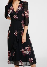 Kvinna klädd i en svart midiklänning med rosa och vita blommönster och trekvarts genomskinliga ärmar, stående med en hand på höften.