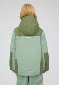 Bambino che indossa una giacca a cappuccio verde a due tonalità e pantaloni abbinati, in piedi di spalle contro uno sfondo semplice.