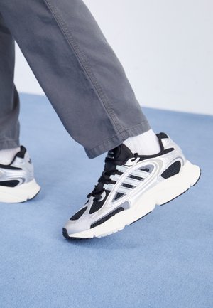 Osoba w szarych spodniach i białych skarpetkach, wykonująca krok do przodu w czarnych i srebrnych sportowych butach na jasnoniebieskiej podłodze.