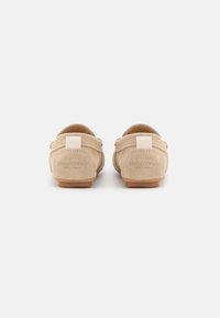 Et par beige ruskinds slip-ons set fra baggrunden med "Marc O’Polo" mærkenavn på hælpanelerne mod en hvid baggrund.