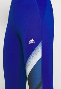 Blå träningsleggings i stretchigt material. Har en vit Adidas-logga och grafiska diagonala mönster i nyanser av blått och svart på sidan.