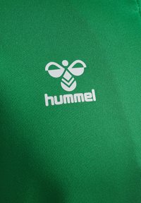 Vêtements de sport verts en tissu texturé, arborant un logo Hummel blanc bien visible, accompagné d'un design stylisé d'abeille et de texte.
