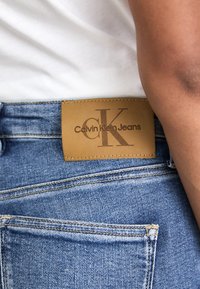 Barna bőr címke "Calvin Klein Jeans" felirattal kék farmernadrágon varrási részletekkel. Oldalán a bőrszín benyomása látható.