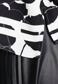 Haut noir et blanc avec des motifs abstraits et des rayures audacieuses, superposé sur un matériau en cuir synthétique noir plissé et texturé.