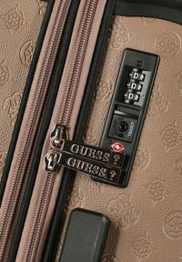 Maleta marrón texturizada con patrones en relieve, cremalleras dobles etiquetadas "GUESS?" bloqueadas en un candado combinado TSA negro ajustado a 0-0-0.