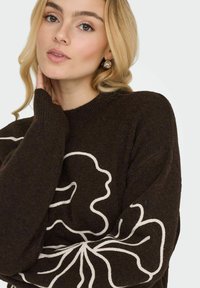 Pull en tricot marron avec un motif floral clair sur le devant, doté de poignets côtelés et d'un col rond.