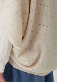 Maglione a maglia beige chiaro con un design a trama aperta, caratterizzato da righe orizzontali e una consistenza morbida, drappeggiato su una camicia bianca a maniche lunghe.