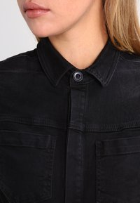 Černá denimová košile s tlačným límečkem, vybavená dvěma předními kapsami a hladkou texturou. Tkanina má jemný lesk.