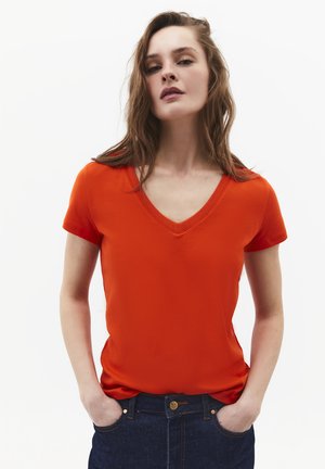 Vrouw met golvend bruin haar die een oranje T-shirt met V-hals en donkerblauwe jeans draagt, handen in de zakken, tegen een effen witte achtergrond.