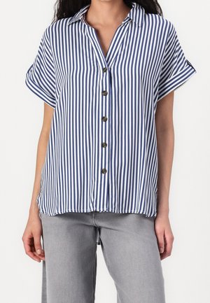 Vrouw draagt een blauw-wit verticaal gestreept overhemd met korte mouwen, gecombineerd met lichtgrijze spijkerbroek.