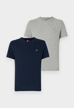 Jeune homme portant un t-shirt bleu marine, un jean beige, des baskets noires et une casquette blanche, debout avec une main dans la poche.