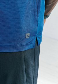 Niebieska techniczna koszulka sportowa z teksturowanej tkaniny i bocznymi rozcięciami, z szarą metką z logo na dolnym boku. Połączona z ciemnymi szortami.