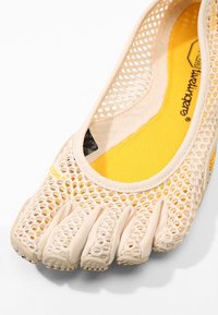 Beige mesh sko med fem tår, en gul innersula, med en flexibel ovandel och gummiyttersula, designad för andningsförmåga och komfort.