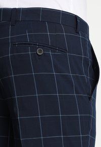 Dunkelblaue Hose mit hellgrauem Fenstergittermuster, ausgestattet mit einer geknöpften Gesäßtasche und einem Gürtelschlaufen am Bund.