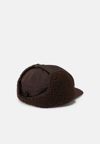 Cappello marrone con un morbido e strutturato copriorecchie in pile, parte superiore liscia e visiera curva. Presenta una cinghia regolabile sul retro.
