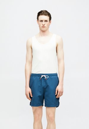 Νέος άνδρας που φοράει λευκή φανέλα και μπλε μαγιό με κορδόνι, στέκεται μπροστά σε απλό ανοιχτό φόντο, κοιτάζοντας μπροστά.